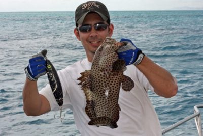 Angler in a fishing cap holds a spotted grouper and topwater lure aboard a Caicos Water Fun charter boat on calm turquoise waters in Turks and Caicos.